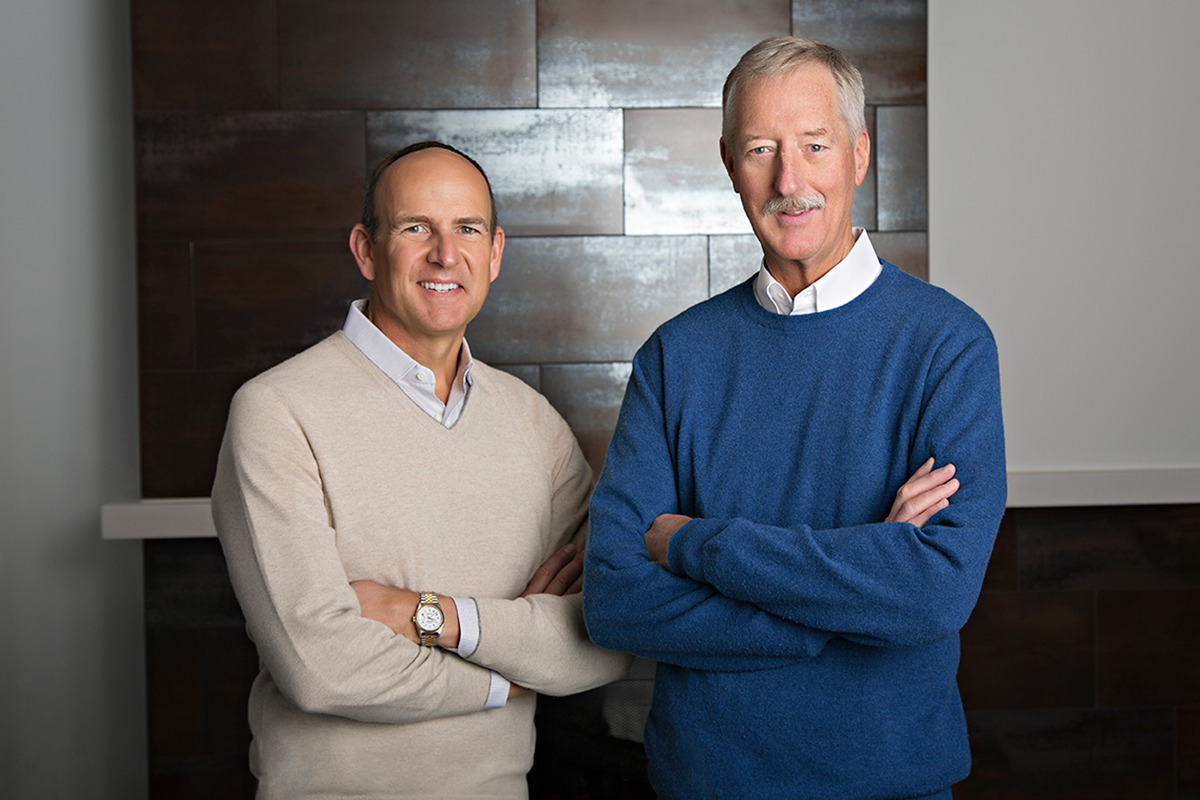 Doug Devos, in white sweater, standing next to Steve Van Andel in a blue sweater, against paneled wall