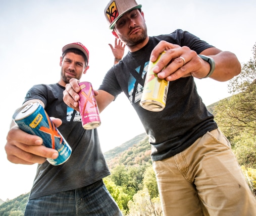 Two men wearing XS shirts and hats hold cans of XS Energy drink towards the camera