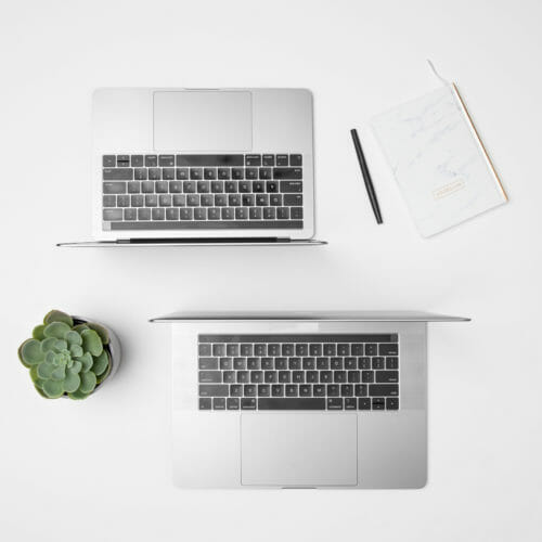 looking down from straight overhead at a clean desk with two open laptops a plant and a notepad and pen