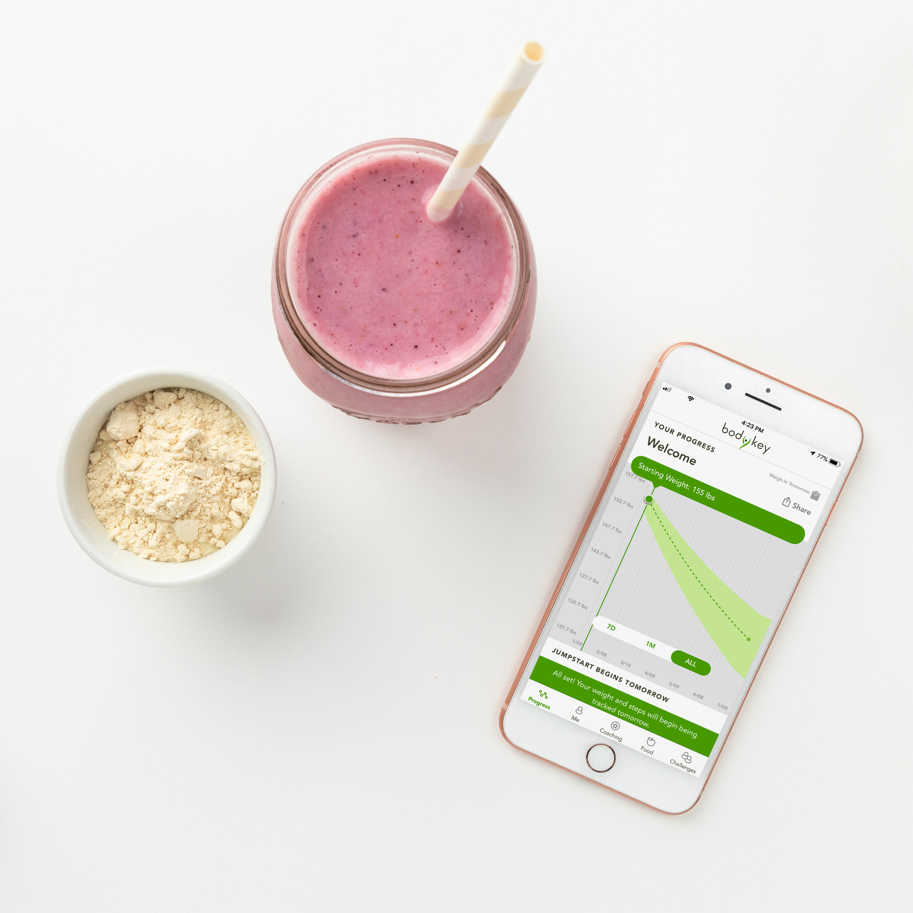 A strawberry BodyKey meal replacement shake in a glass with a straw is placed between a ramekin of the vanilla shake mix and a smartphone linked to the BodyKey Digital Coach. The Digital Coach allows you to stay connected, engaged and motivated on your terms.