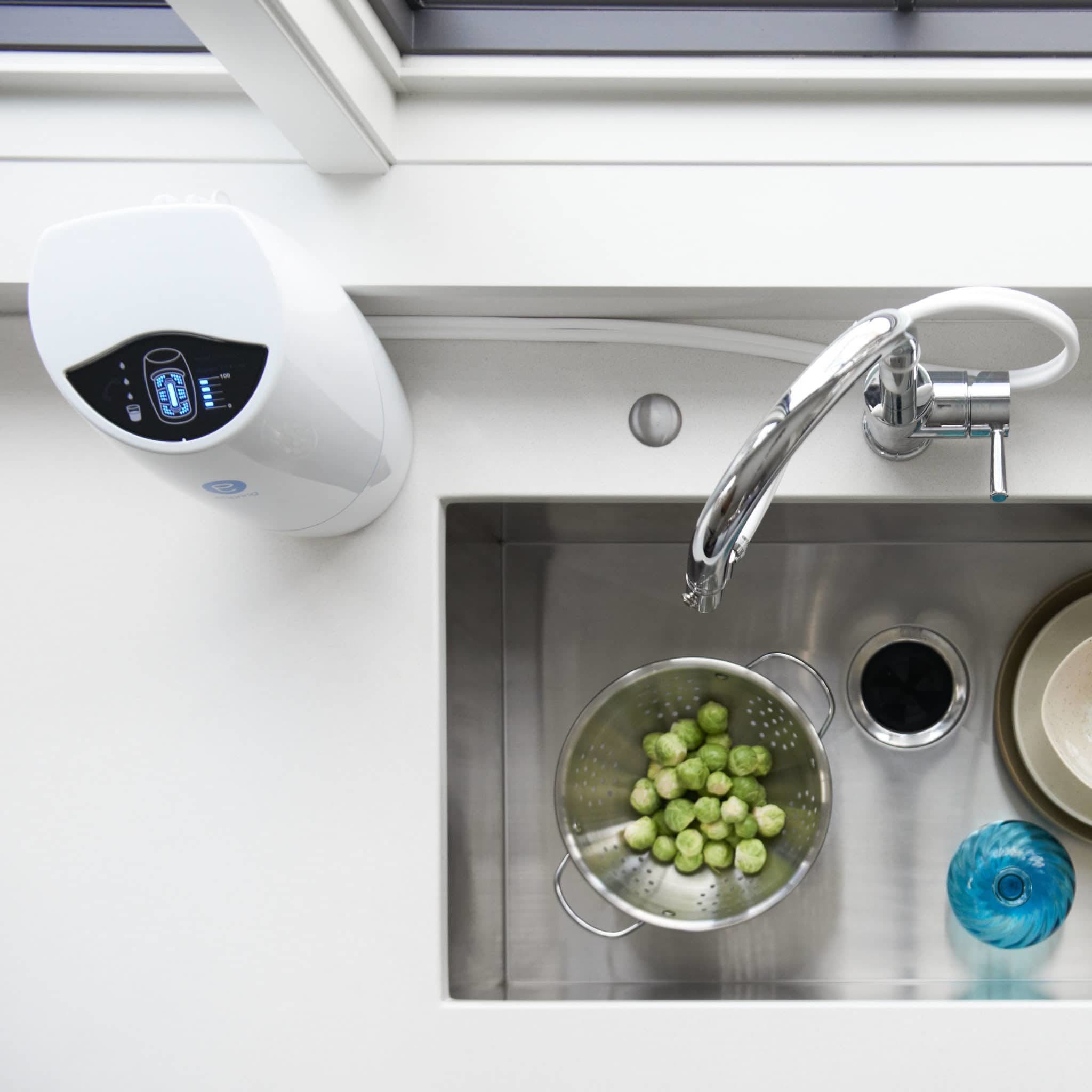 Overhead view of a kitchen sink connected to an eSpring water treatment system. A colander of brussels sprouts, a glass vase and plates are arranged in the sink. The eSpring's pressed carbon block filter effectively removes 140+ potential health-effect contaminants that may be present in drinking water, including lead, mercury, disinfectant by-products and volatile organic compounds.