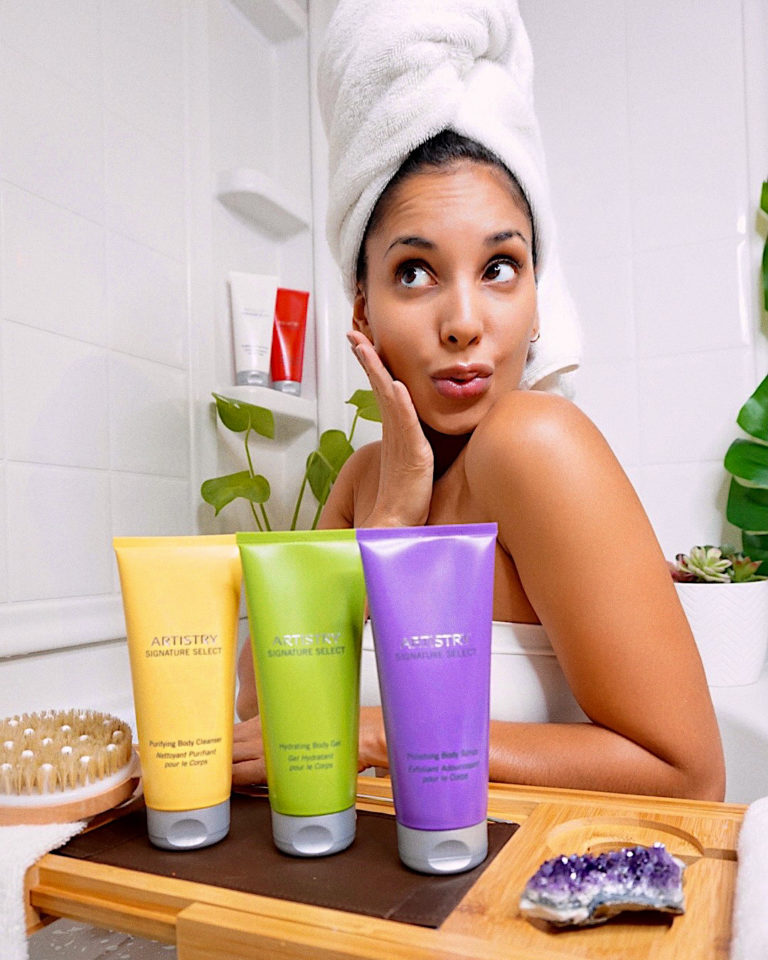 Woman sitting in a bathtub with a towel on her head posing with artistry studio select products in front of her on a bath tray and some more behind her on the shower shelf