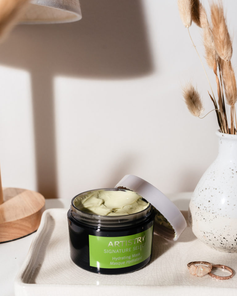 Artistry Signature Hydrating Mask sitting on a white dish with a vase with wheat to its right. There is a candle in the left background and rings in the dish.