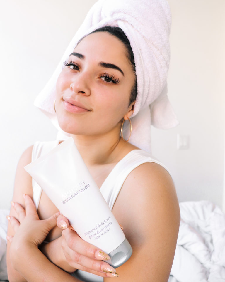 Woman with a towel on her head giving herself a hud holding a bottle of artistry signature select brightening body cream