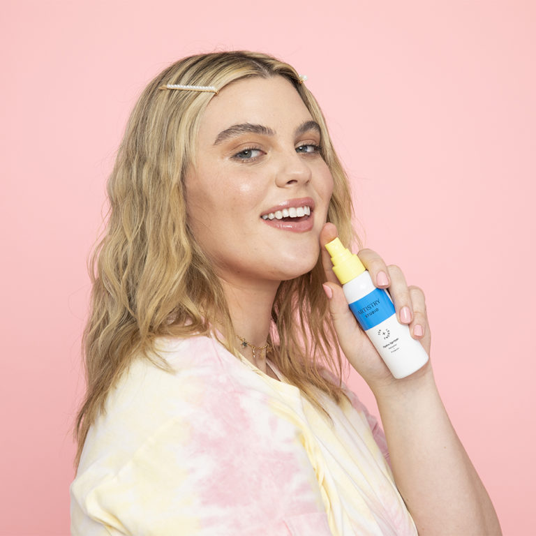 Woman standing in front of a pink backdrop smiling holding a spray bottle of Artistry studio hydro-spritzer up to her face.