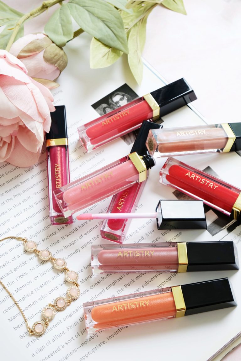 Eight different colored artistry lip glosses sitting on top of a book with a necklace and peony sitting next to them