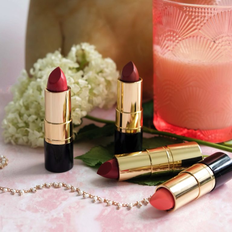 Four different colored lipsticks without the cap on sitting on a pink surface with hydrangeas, a candle, and a necklace around it.