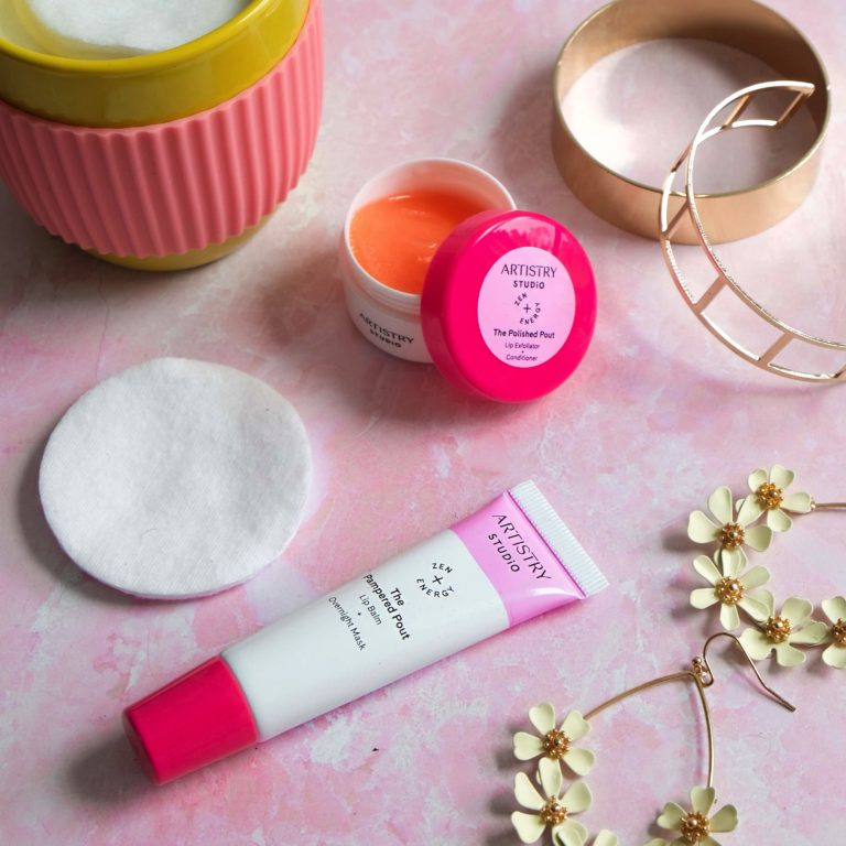 Artistry studio the polished pout and the pampered pout sitting on a pink counter with a candle, earrings, bracelets, and a cotton swab.