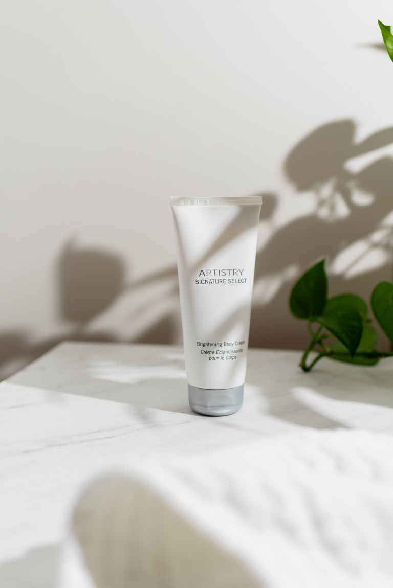 Bottle of Artistry brighting body cream sitting on a marble surface with a plant next it.