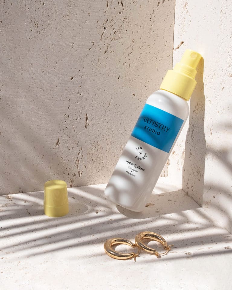 bottle of artistry studio hydro-spritzer leaning against a wall with a pair of gold earrings sitting in front of it